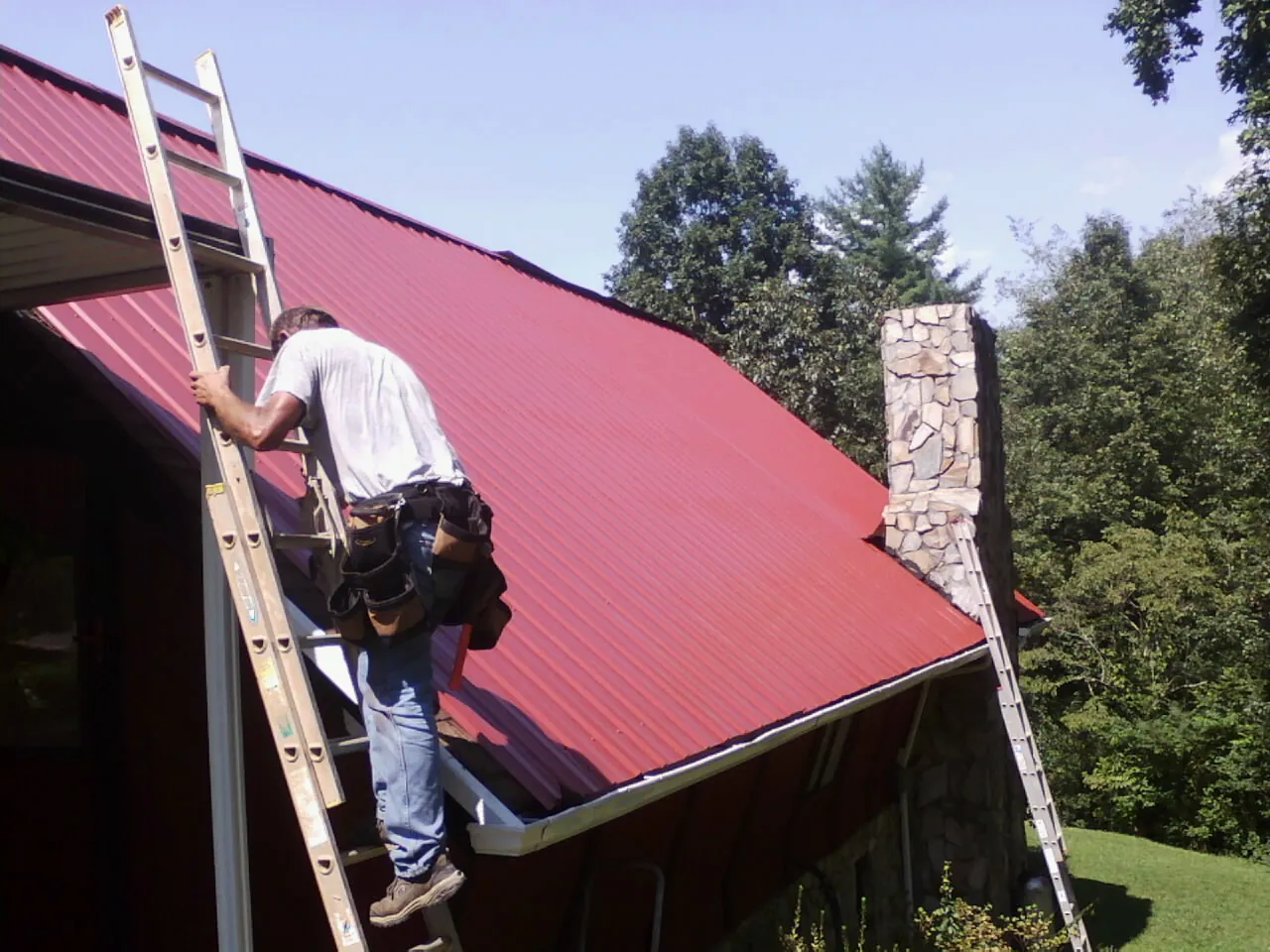 Roofer on a ladder beside a red metal roof and stone chimney — Soffit & Fascia Repair by Roofing Brothers in Shamokin Dam